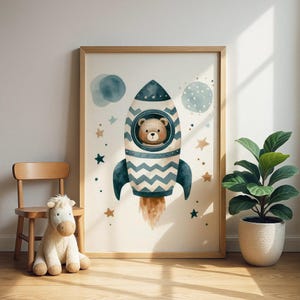 May include: A framed watercolour print depicts a teddy bear in a rocket ship. The rocket is white and blue with a zig-zag design. The background includes stars, planets, and a light beige backdrop. The frame is made of light brown wood.