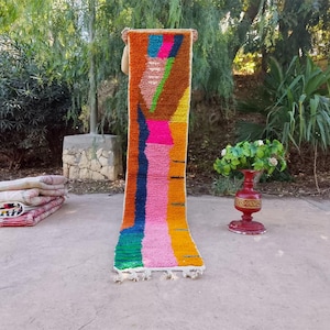 Op de afbeelding: Een kleurrijk, rechthoekig tapijt met een levendig abstract ontwerp. Het tapijt heeft een mix van oranje, roze, blauwe, groene en gele tinten. Het tapijt wordt gepresenteerd tegen een natuurlijke buitenachtergrond.