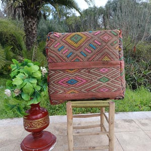 Puede incluir: Un cojín cuadrado estampado con un diseño geométrico en tonos rosa, amarillo, azul y verde. El cojín está colocado sobre un pequeño taburete de madera. Un jarrón rojo con un arreglo floral está en primer plano.