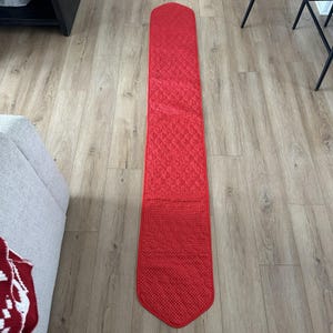 May include: A long, rectangular red table runner with a quilted diamond pattern. The bottom portion has a dotted design. The runner is displayed on a light wood-grain floor.