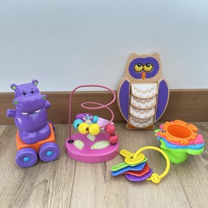 Pode incluir: Uma coleção de brinquedos coloridos para bebês, incluindo um hipopótamo roxo sobre rodas, um labirinto de contas rosa, uma coruja de madeira, uma flor empilhável e um conjunto de chaves coloridas. Projetado para o desenvolvimento infantil.