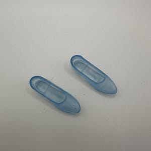 May include: Two small, translucent blue ballet flats. The shoes are made of a soft plastic material and have a rounded toe. The shoes are lying on a white surface.