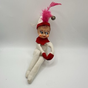May include: A vintage elf doll with a white body, red mittens, and a red collar. The doll has a painted face, a white and red hat with a pink feather, and a small gold bell.