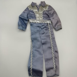 May include: A two-piece doll outfit in shades of blue and gray. The top has a silver sequined collar and chest detail, with a matching sequined belt. The pants have silver trim down the sides. The outfit is made of a shiny, satin-like material.