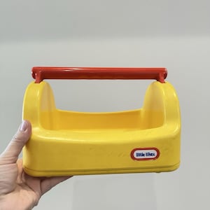 May include: A yellow plastic toy toolbox with a red handle. The toolbox has rounded edges and a rectangular shape. The words "little tikes" are printed on the side of the toolbox.