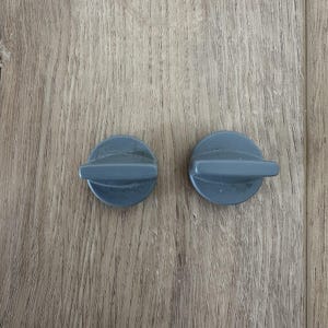 May include: Two gray, circular knobs with a horizontal, rectangular handle extending from the center. The knobs are positioned on a light brown, wood-grain surface.
