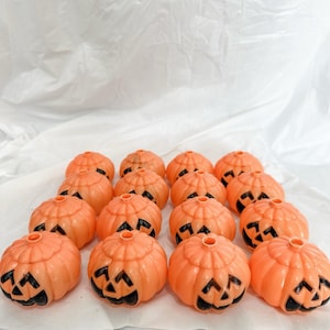 May include: A collection of orange plastic jack-o'-lanterns with black cut-out faces. The pumpkins are arranged on a white surface. Each pumpkin has a stem and a carved face, perfect for Halloween decorations.