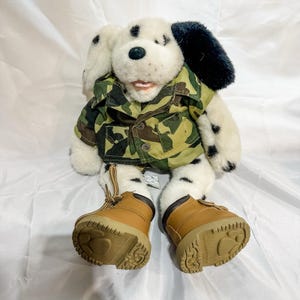 May include: A plush Dalmatian dog wearing a camouflage jacket and brown boots. The dog has black spots, black ears, and a pink tongue. The boots have brown laces and soles.