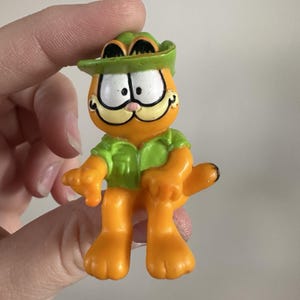 May include: A small, orange Garfield figurine wearing a green safari hat and shirt. The figurine has white eyes, a pink nose, and a black-outlined mouth. The figure is holding a pose with its arms and legs extended.