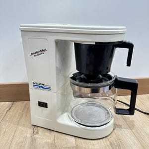 May include: A white Proctor Silex coffee maker with a black filter basket and a glass carafe. The coffee maker has a power switch and the text "Proctor Silex 2 to 12 CUPS" is visible on the side. The carafe has a black handle.