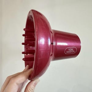 May include: A burgundy-colored Vidal Sassoon hair dryer diffuser. The diffuser has a round, bowl-like shape with numerous finger-like extensions. The brand name is printed on the side of the diffuser.