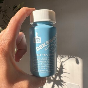 Può includere: Una bottiglia blu chiaro di Dekswood Exterior Wood Cleaner con tappo bianco. La bottiglia presenta il marchio "Flood" e i dettagli del prodotto stampati sull'etichetta. È visibile anche il testo "Sold Thru Paints".