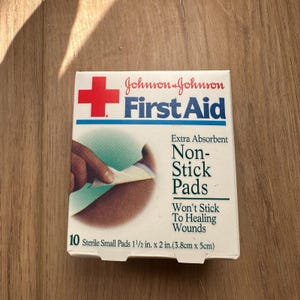 Puede incluir: Una caja blanca de apósitos First Aid Non-Stick de Johnson & Johnson. La caja presenta una cruz roja y el texto "Extra Absorbent" y "Won't Stick To Healing Wounds". La caja contiene 10 apósitos pequeños estériles, cada uno de 3,8 cm x 5 cm.
