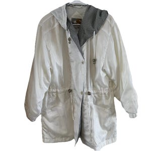May include: A white, hooded jacket with a drawstring waist and button closures. The jacket features a black and white checkered pattern lining the hood and interior. The jacket has long sleeves and a relaxed fit, suitable for outerwear.