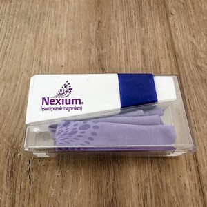 Puede incluir: Una caja blanca y azul que contiene un artículo morado y un objeto rectangular blanco y azul. La caja tiene la palabra "Nexium" impresa en morado. El artículo dentro de la caja es de color morado claro.