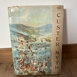 May include: A vintage book titled "The Custer Myth" by Graham. The cover features a detailed illustration of a battle scene with soldiers on horseback, set against a backdrop of rolling hills and a cloudy sky. The book's spine is a light beige color.