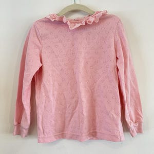 Pode incluir: Uma camisa rosa claro de manga comprida com um padrão delicado em forma de coração. A camisa apresenta uma gola e punhos com babados. O tecido tem uma textura subtil e a camisa está pendurada num cabide de madeira.