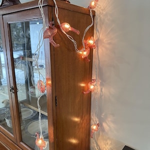May include: A string of pink flamingo-shaped lights with a white wire. The lights are illuminated, casting a warm glow. The string is draped against a wooden cabinet.