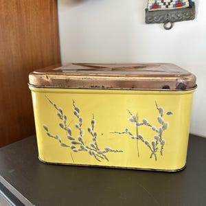 Op de afbeelding: Een rechthoekige gele blikken doos met een koperkleurig deksel. De doos is versierd met een bloemmotief van witte en grijze katjes. De doos is waarschijnlijk vintage en zou geschikt zijn voor opslag.