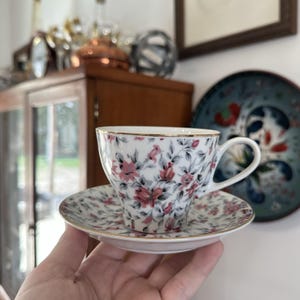 May include: A white teacup and saucer set with a floral pattern in pink, gray, and gold. The teacup and saucer have a gold trim. The teacup is sitting on the saucer. The background includes a wooden cabinet and a decorative plate.
