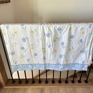 Puede incluir: Sábana floral blanca con un estampado de flores amarillas y azules y hojas verdes. La sábana tiene un borde azul claro con flores blancas. La sábana está sobre una barandilla de madera.