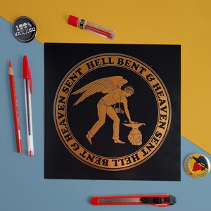May include: A black and gold print with a circular design featuring a winged figure holding a hammer and anvil. The text "SENT HELL BENT & HEAVEN SENT HELL BENT & HEAVEN" is written around the circle.
