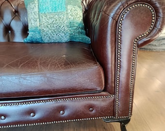 Puede incluir: Un sofá Chesterfield de cuero marrón con respaldo y reposabrazos capitoné. Un cojín cuadrado decorativo con un diseño de patchwork en tonos turquesa y beige descansa sobre el asiento. El sofá tiene ribete de tachuelas y patas de madera oscura.