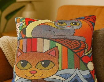 May include: Decorative square pillow featuring a colorful cat design. The pillow has a brown cat at the bottom and a gray cat at the top, with a vibrant mix of colors including red, orange, and blue. The pillow is placed on an orange couch.
