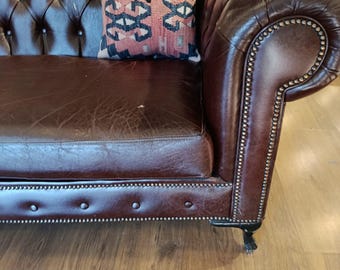 May include: A brown leather Chesterfield sofa with a patterned pillow. The sofa has a tufted back and armrests with nailhead trim. The pillow has a geometric design in shades of red, blue, and cream.