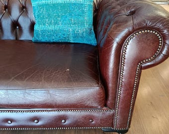 Puede incluir: Un cojín con estampado turquesa y blanco descansa sobre un sofá Chesterfield de cuero marrón. El sofá tiene un diseño capitoné y ribete con tachuelas. El fondo presenta una pared de ladrillo y un suelo de madera.