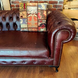 May include: A tufted, burgundy leather sofa with a decorative pillow. The pillow features a patchwork design with various colors and patterns. The sofa has a rolled arm and nailhead trim.