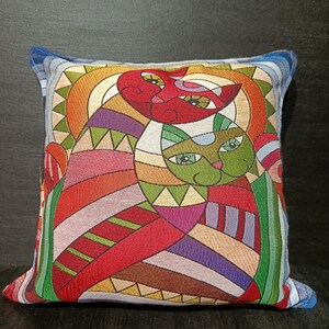 May include: Square decorative pillow featuring a colorful abstract design of two cats. The cats are depicted in red and green with geometric patterns and a sun-like background. The pillow has a blue border.