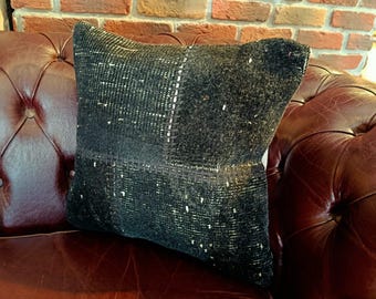 Puede incluir: Un cojín cuadrado gris oscuro con un diseño tejido y texturizado. El cojín tiene una apariencia de patchwork con detalles blancos y está colocado sobre un sofá de cuero marrón. El fondo presenta una pared de ladrillo.
