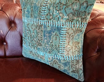Puede incluir: Un cojín cuadrado turquesa con un diseño de patchwork. El cojín presenta una mezcla de patrones en tonos de azul, verde y gris. Se coloca sobre un sofá de cuero marrón, creando un contraste de texturas y colores.