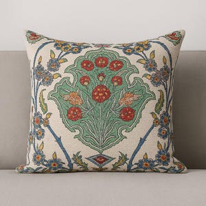 Op de afbeelding: Vierkant decoratief kussen met bloemenpatroon. Het crèmekleurige kussen heeft een centraal groen bloemmotief met rode bloemen en blauwe en gele accenten. Het ligt op een grijze bank.