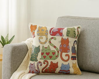May include: Square throw pillow with a cream background featuring a colorful pattern of cartoon cats. The cats are in various poses and colors, including green, orange, and blue, with heart and floral details.