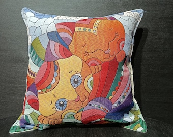 May include: Square decorative pillow featuring a colorful illustration of two cartoon cats. The cats are depicted in shades of orange, yellow, red, and blue, with intricate patterns and details. The pillow has a white and blue patterned border.