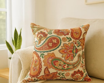 May include: A square decorative pillow with a colorful paisley pattern in shades of orange, red, green, and teal on a cream background. The pillow is placed on a beige sofa with a cream-colored throw blanket.