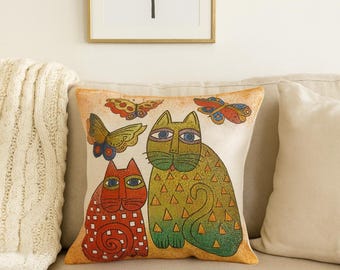 May include: Decorative throw pillow featuring a whimsical design of two cats and butterflies. The cats are in shades of green and red, with patterned details. The pillow is on a beige sofa, with a framed palm tree print in the background.
