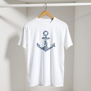 Puede incluir: Camiseta blanca con un dise&ntilde;o de ancla azul. El ancla est&aacute; compuesta por patrones n&aacute;uticos en azul y blanco. La camiseta cuelga de una percha de madera sobre un fondo blanco.
