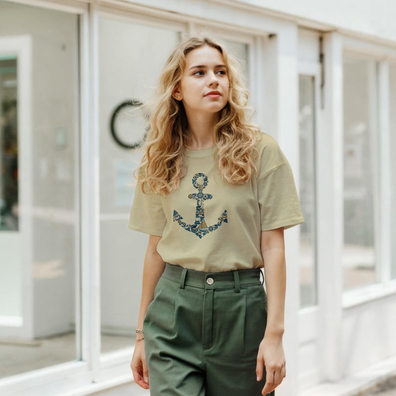 Puede incluir: Una camiseta beige con un dise&ntilde;o de ancla azul marino y blanco. El ancla est&aacute; compuesta por elementos florales y n&aacute;uticos. La persona lleva pantalones verdes. El fondo es un edificio blanco.
