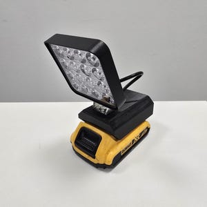 May include: A black LED work light attached to a yellow and black DeWalt battery. The light has a rectangular head with multiple LED bulbs. The battery is a standard size for power tools.