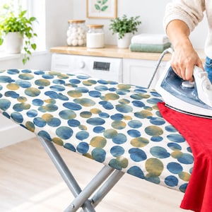 May include: An ironing board cover featuring a white background with a repeating pattern of overlapping blue and gold watercolour-style circles. The board is supported by a grey metal frame, and a white and grey steam iron is pressing a red garment.