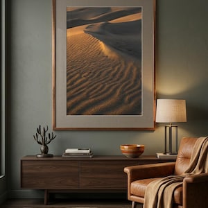 Può includere: Una fotografia verticale incorniciata di dune di sabbia con luce dorata e ombre profonde, esposta sopra una console in legno scuro. Il tavolo ospita una scultura scura, libri impilati e una ciotola, accanto a una poltrona in pelle.