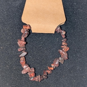May include: A bracelet composed of small, irregularly shaped, reddish-brown gemstones. The bracelet is strung together and presented on a brown tag with a small hole for hanging. The background is a dark, textured surface.