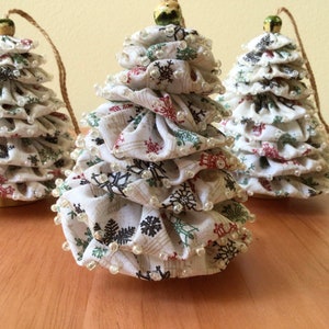 May include: Three handmade Christmas tree ornaments. Each tree is made of white fabric with red, green, and black snowflake patterns. The trees are adorned with small, clear beads and have a wooden base and a twine loop for hanging.