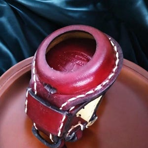 Pode incluir: Um item de couro bordô feito à mão com costura branca. Possui uma abertura circular e uma tira com um detalhe dourado. O couro parece envelhecido, com sinais visíveis de desgaste.