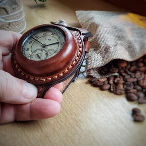 Puede incluir: Un reloj de bolsillo vintage con una funda de cuero marrón y detalles cosidos. La esfera del reloj tiene agujas negras y una subesfera. El reloj se sostiene en una mano, con granos de café y un saco de yute en el fondo.