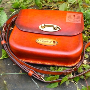 Peut inclure: Sac bandoulière en cuir orange fait à la main, avec fermoir en laiton et bandoulière réglable. Le sac a une forme arrondie et une petite plaque signalétique en laiton avec le nom "Grakoff". Édition limitée.
