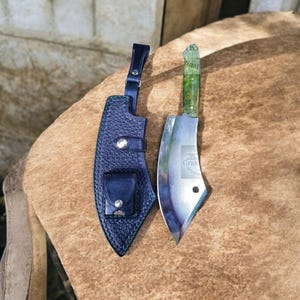 Puede incluir: Un cuchillo de carnicero con mango verde y hoja plateada, junto a una funda de cuero azul oscuro. El cuchillo tiene el texto "Grak Original" en la hoja. La funda tiene un pequeño bolsillo y una correa.